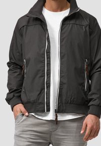 Veste bomber noire avec fermeture éclair à l'avant, poignets et col côtelés. Comprend deux poches zippées avec tirettes en cuir marron. Tissu léger.