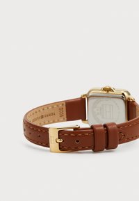 Reloj con correa de cuero marrón y caja cuadrada en tono dorado, con acentos cosidos y un fondo transparente que muestra el logo y la información.