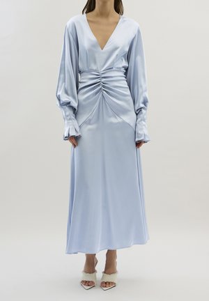 Kvinde iført en lyseblå satin kjole med lange ærmer, rynket talje og dyb V-udskæring, kombineret med hvide spidse hæle med pelskant.