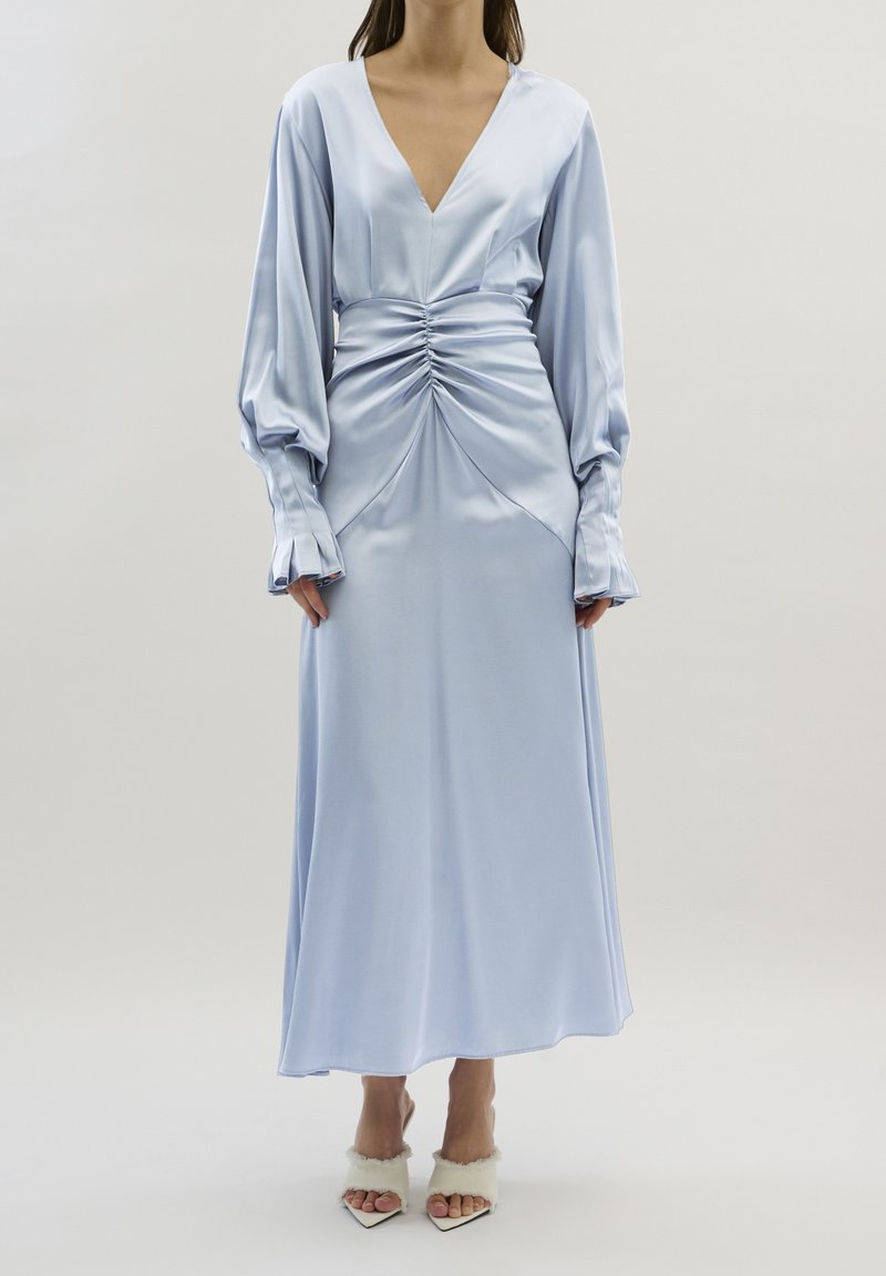 Femme portant une robe bleu clair en satin à manches longues avec une taille froncée et un décolleté en V profond, associée à des talons pointus blancs bordés de fourrure.