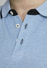 Lichtblauwe polo met een textuurstof, zwarte kraag en witte knopen. Bevat zwarte geborduurde accenten bij de knopen.