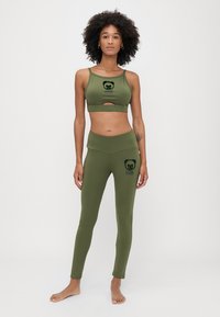 Sport brassiere i tajice u maslinastoj boji, izrađeni od rastezljivog materijala. Brassiere ima dizajn s izrezom i logotip medvjeda; tajice imaju odgovarajući logotip i visok struk.