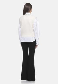 Gilet bianco a maglia con orlo a coste e dettagli decorativi sul colletto, indossato sopra una camicia bianca e pantaloni svasati neri.