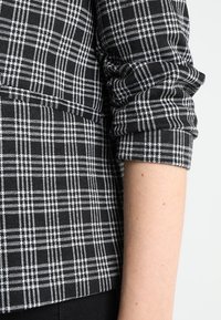 Černo-bílá kostkovaná košile s krátkým rukávem a strukturovanou látkou, s ohrnutými rukávy a tvarovaným lemem. Doplňky zahrnují ostré linie.