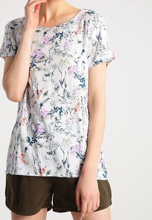 Femme portant une chemise blanche à manches courtes avec un motif floral coloré et un short vert olive foncé sur un fond uni.