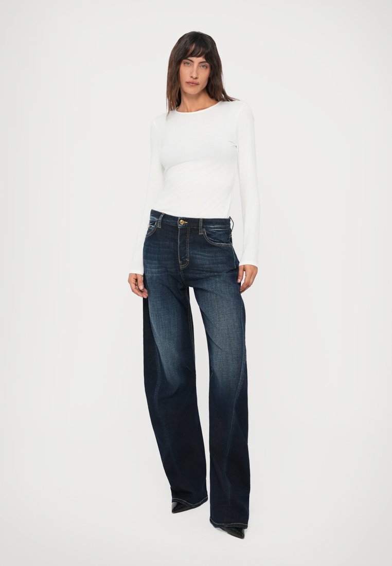 Femme portant un haut blanc ajusté à manches longues et un jean bleu foncé délavé à jambes larges, se tenant devant un fond blanc uni.