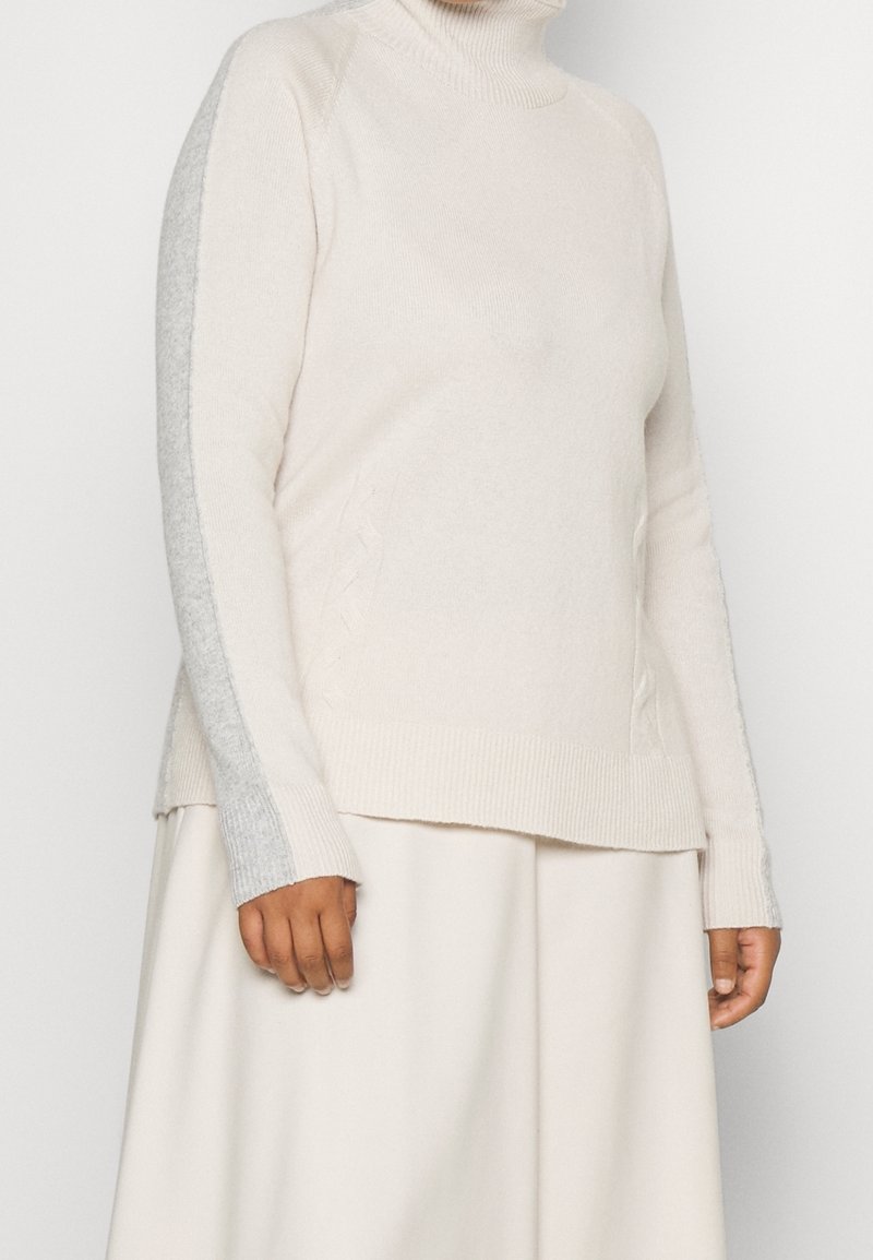 Femme portant un pull à col roulé beige clair avec des rayures grises sur les manches et une jupe crème assortie.