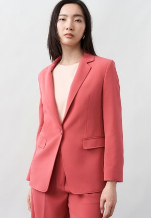 Femeie purtând un sacou roz coral și pantaloni asortați peste un top bej deschis, stând în picioare pe un fundal simplu și deschis la culoare.