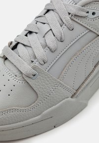 Primer plano de una zapatilla de color gris claro que muestra cuero texturizado, cordones trenzados, costuras detalladas y parte de la suela de goma.