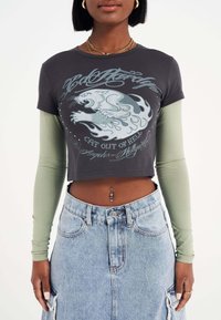 Dunkelgraues, cropped T-Shirt mit grafischem Löwenmotiv, über einem langen, olivgrünen Oberteil getragen. Kombiniert mit einem hoch geschnittenen, hellblauen Denim-Rock.