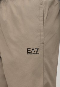 Μπεζ παντελόνι με κάθετη πλαϊνή τσέπη και μαύρο λογότυπο "EA7 Emporio Armani" τυπωμένο κοντά στην τσέπη.