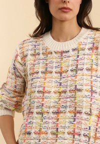 Femme portant un pull en tricot crème avec des motifs de fil multicolores verticaux et horizontaux, ainsi que des poignets et un col côtelés.