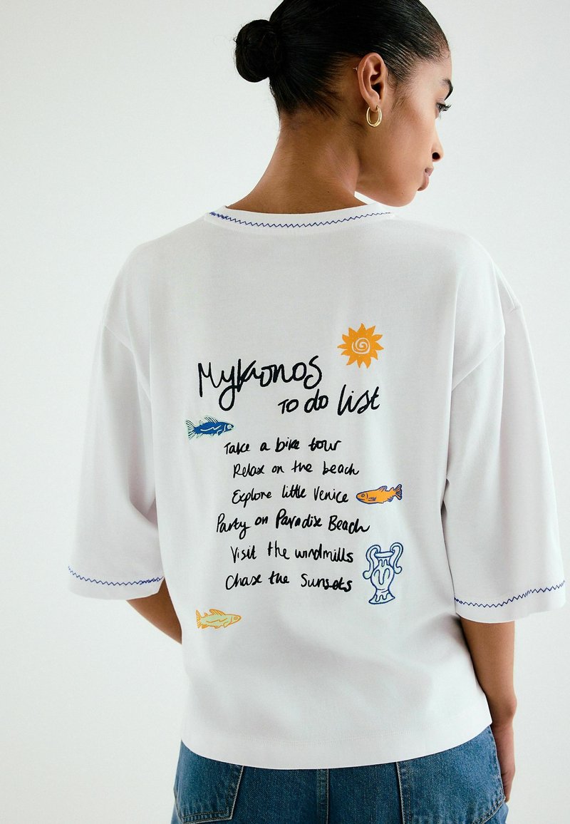 Femme aux cheveux attachés en chignon portant un T-shirt blanc avec la mention "Mykonos to do list" et des illustrations colorées de poissons, de soleil et de vase dans le dos.