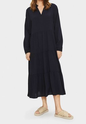 Granatowa sukienka na długi rękaw z dekoltem w serek, wielowarstwowym fasonem i marszczoną talią. Noszona z jasnobeżowymi sandałami wsuwanymi.