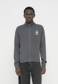 Γκρι πλεκτό πουλόβερ με φερμουάρ και ριμπ ύφανση, με ψηλό γιακά, με κεντημένο κρατικό έμβλημα στο στήθος και εφαρμοστά μανικάκια. Υφή με πλεκτό σχέδιο.