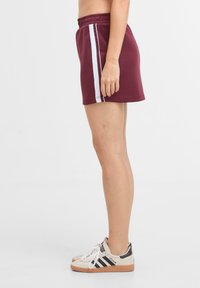 Shorts sportivi bordeaux con strisce bianche sui lati, cintura elastica e vestibilità rilassata, abbinati a scarpe da ginnastica beige con dettagli neri.