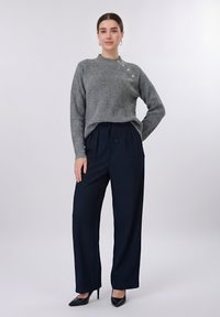 Γυναίκα που φοράει γκρι πλεκτό πουλόβερ με ασημένια κουμπιά και φαρδιά μαύρη παντελόνα, στέκεται με το ένα χέρι στη μέση.