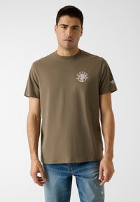 Braunes Baumwoll-T-Shirt mit Rundhalsausschnitt, vorne auf der Brust ein rundes Logo in Weiß und Rot sowie eine kleine Grafik am Ärmel.