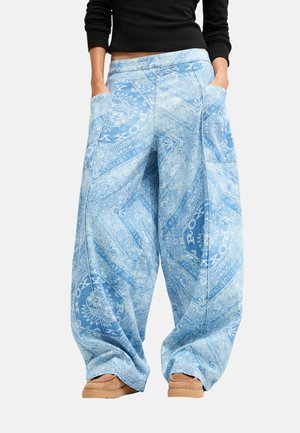 Personne portant un pantalon large bleu clair avec des motifs blancs complexes et des poches, associé à des chaussures marron clair et un haut noir à manches longues.