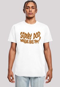 Bílý bavlněný tričko s výrazným hnědým textem, který říká "SCOOBY DOO KDE JSI!" ve hravém, nepravidelném písmu.