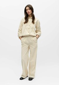 Camisa beige abotonada con bolsillo y pantalones beige de corte holgado, con superficie texturizada y cintura elástica, combinados con zapatos negros.