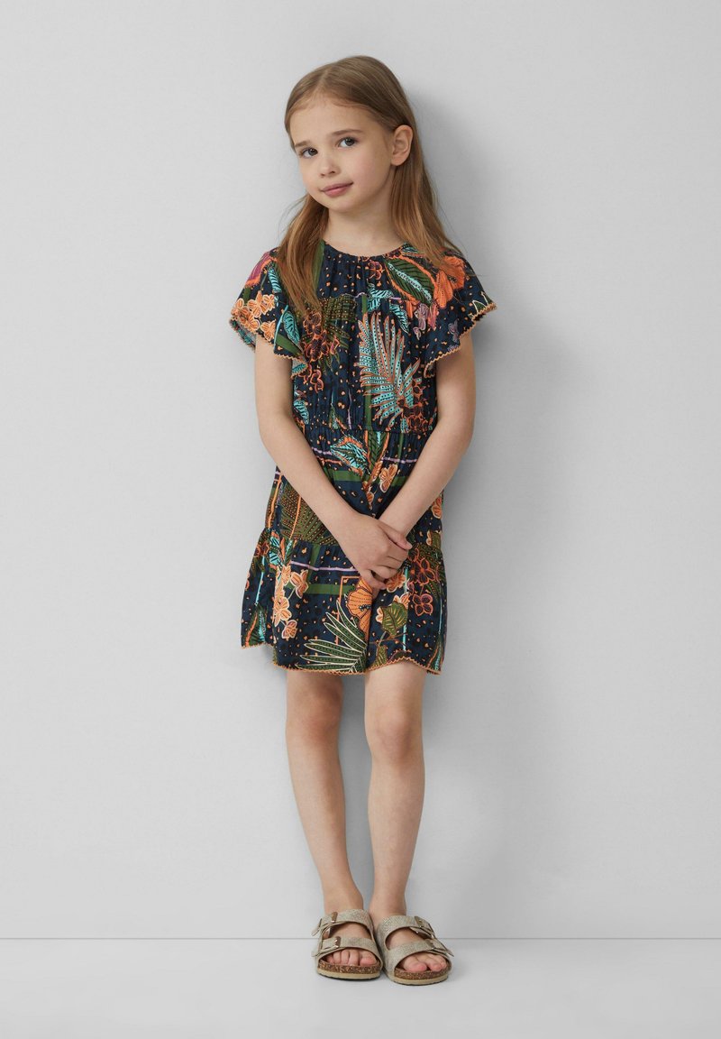 Blumenmuster-Kleid mit Rüschensäumen, dunkelblauem Hintergrund und leuchtenden orange und grünen Akzenten. Kombiniert mit hellbeigen Sandalen.