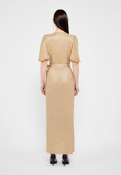 Rochie maxi aurie, cu mâneci lungi, cu o siluetă mulată, dintr-un material texturat, cu mâneci bufante și o curea legată în talie. Sandale cu toc înalt negre vizibile.