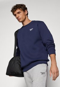 Granatowy sweatshirt Reebok z okrągłym dekoltem i ściągaczami, zestawiony z szarymi spodniami dresowymi, z logo Reebok i czarną torbą na ramię.