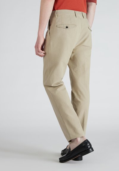 Beżowe chinosy o prostym kroju, z wykończeniem tylnych kieszeni, gładką fakturą, połączone z czarnymi loafersami na jasnym tle.