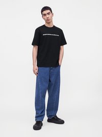Νέος άνδρας που στέκεται, φοράει ένα μαύρο t-shirt Emporio Armani, φαρδιά μπλε τζιν και μαύρα αθλητικά παπούτσια μπροστά σε απλό φωτεινό φόντο.