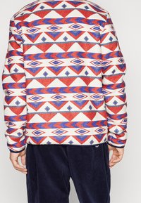Multikleurige sweatshirt met een geometrisch patroon van rode, blauwe en witte diamanten op een gestructureerde stof; gecombineerd met marineblauwe corduroy broeken.
