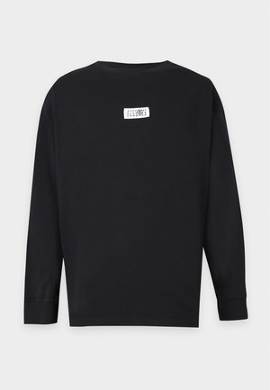 Joven hombre con gorra negra, camisa negra de manga larga con pequeño logo blanco en el pecho, pantalones negros holgados y zapatos negros, de pie frente a fondo blanco.