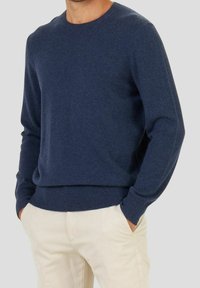Hombre con suéter de cuello redondo azul marino y pantalones beige, de pie con las manos en los bolsillos frente a un fondo liso.