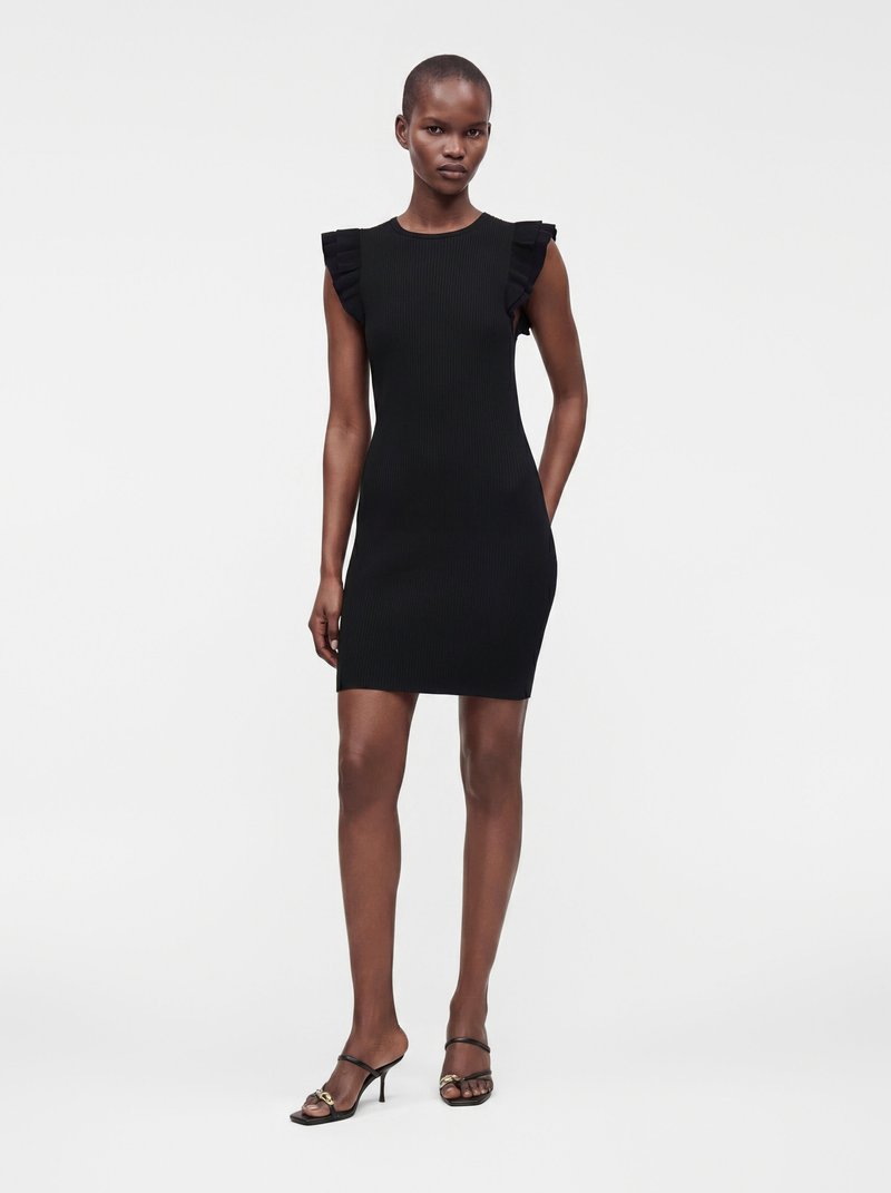 Femme debout portant une robe noire sans manches côtelée avec des épaules à volants et des sandales à talons noires à lanières sur un fond uni.