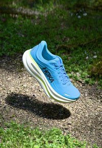 Sapatilha de corrida Hoka azul no ar acima de um caminho de gravilha, com erva verde e pequenas flores ao fundo.