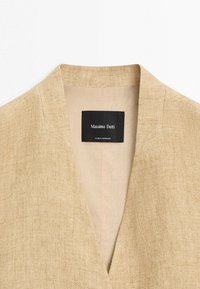 Béžová texturovaná látková bunda s hlubokým výstřihem do V a černou štítkem uvnitř s nápisem "Massimo Dutti, Made in Morocco."