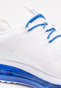 Vita sneakers med en texturerad mesh-övre, blå snören och en blå dämpad sula. Har cirkulära perforeringar för andningsförmåga.