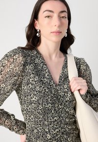 Robe noire à fleurs avec des manches bouffantes transparentes, un corsage à volants et un décolleté en V profond. Un modèle tient un sac à bandoulière crème, agrémenté de grandes boucles d'oreilles.
