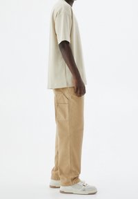 Mężczyzna w beżowej, oversize'owej koszulce, jasnobrązowych prostych spodniach z bocznymi kieszeniami oraz białych trampkach, stojący na białym tle.