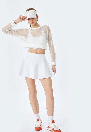 Top corto de malla blanca de manga larga, falda blanca ajustada con dobladillo acampanado, combinada con zapatillas blancas con acentos naranjas y calcetines cortos.