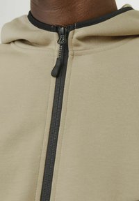 Béžová mikina na zips s čiernym zipsom a lemom. Tkanina sa javí ako hladká a jemná, disponuje kapucňou a priliehavým strihom.