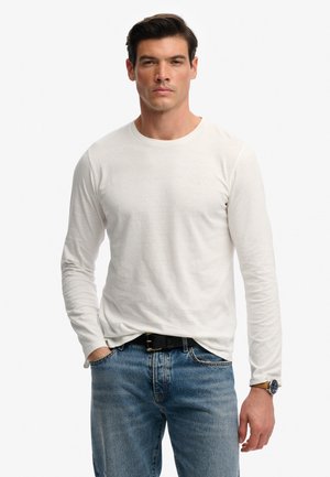 Uomo con camicia bianca a maniche lunghe, jeans blu, cintura nera e orologio da polso, in piedi con una mano in tasca, sfondo neutro.