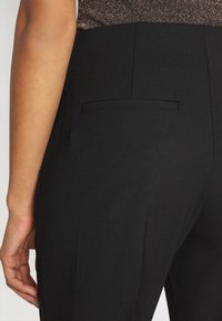 Svarta skräddarsydda byxor med en slät yta, utrustade med en enda bakficka med en dold öppning och utan synlig hårdvara.