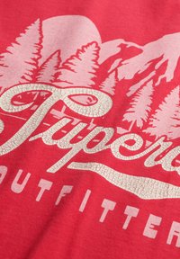 Czerwona bawełniana koszulka z białym popękanym napisem "Supreme Outfitters" oraz wzorem gór i drzew sosnowych w tle.