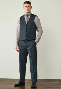 Tummansininen räätälöity puku, jossa on liivi, teksturoitu kangas, kuusi nappia ja slim-fit-housut. Harmaa paita ja kuvioitu solmio. Mustat kengät.