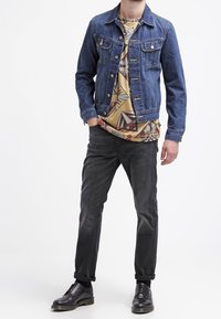 Jeansjacka i blått med metallknappar, mönstrad beige skjorta, svarta jeans och svarta snörstövlar. Avslappnad lager-på-lager-outfit.