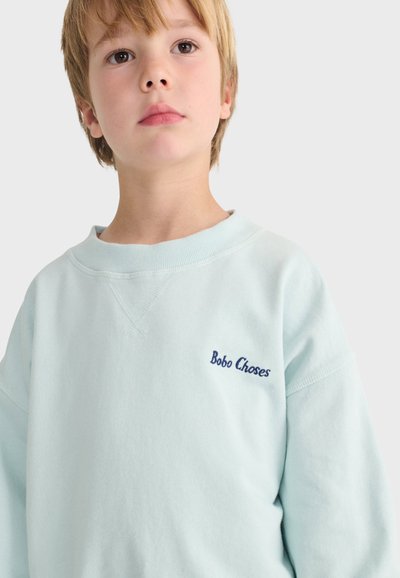Jeune enfant aux cheveux blonds portant un sweat-shirt bleu clair avec "Bobo Choses" brodé sur la poitrine, fond clair neutre.