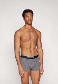 Gray boxer briefs with black accents and Lacoste branding. Features small embroidered logos, close-fitting design, and cotton material.