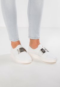 Vita slip-on skor har en slät läderöverdel med ett metalliskt band som visar logotypen. Sulan är tjock och rundad, vilket ger stöd.