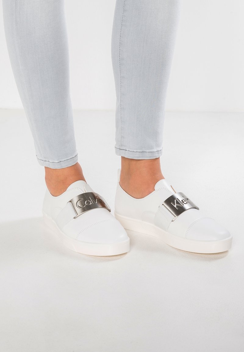 Vita slip-on skor har en slät läderöverdel med ett metalliskt band som visar logotypen. Sulan är tjock och rundad, vilket ger stöd.